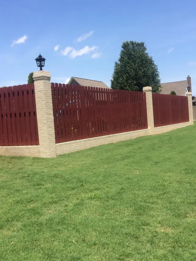 Dark board fence between tan brick columns with lamp caps at a driveway entry